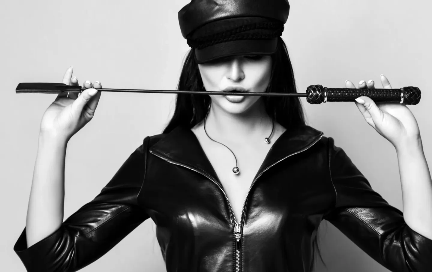 Woman in leather outfit holding a whip across her face.